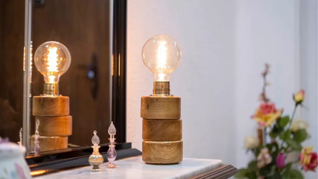 Lámpara Lasai encendida, con base de cilindros de madera de roble dispuestos a desnivel, sobre una cómoda frente a un espejo, con reflejo y decoración floral.