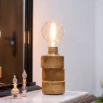 Lámpara Lasai encendida, con base de cilindros de madera de roble dispuestos a desnivel, sobre una cómoda frente a un espejo, con reflejo y decoración floral.