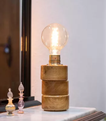 Lámpara Lasai encendida, con base de cilindros de madera de roble dispuestos a desnivel, sobre una cómoda frente a un espejo, con reflejo y decoración floral.