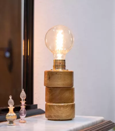 Lámpara Lasai encendida, con base de cilindros de madera de roble dispuestos a desnivel, sobre una cómoda frente a un espejo, con reflejo y decoración floral.