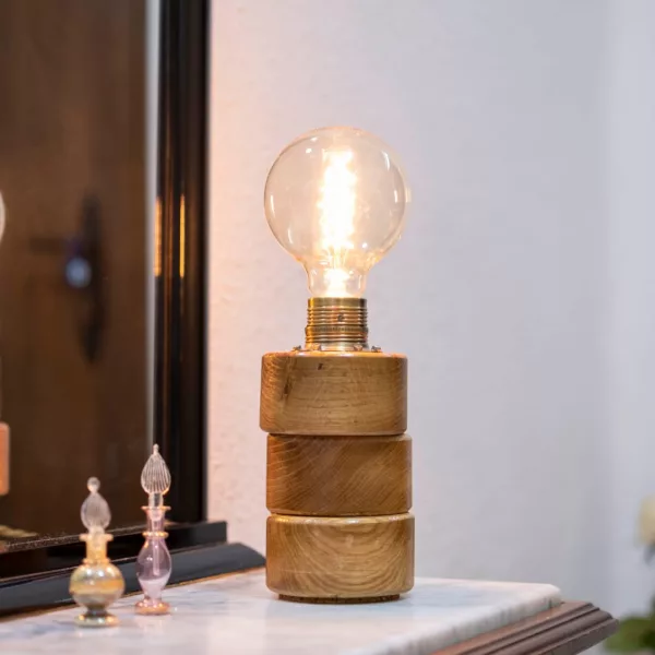 Lámpara Lasai encendida, con base de cilindros de madera de roble dispuestos a desnivel, sobre una cómoda frente a un espejo, con reflejo y decoración floral.