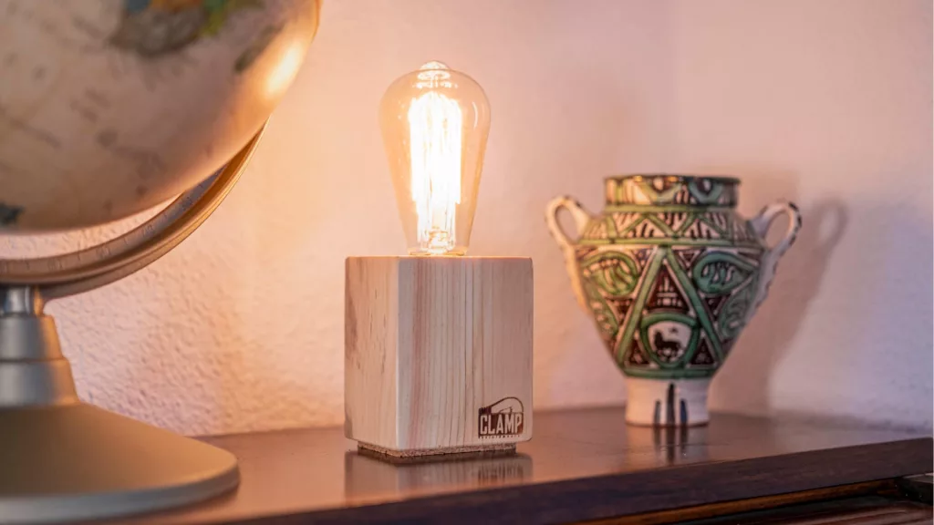 Lámpara Nara encendida, con base cúbica de madera clara, sobre una estantería junto a un globo terráqueo y una pieza de cerámica artesanal.