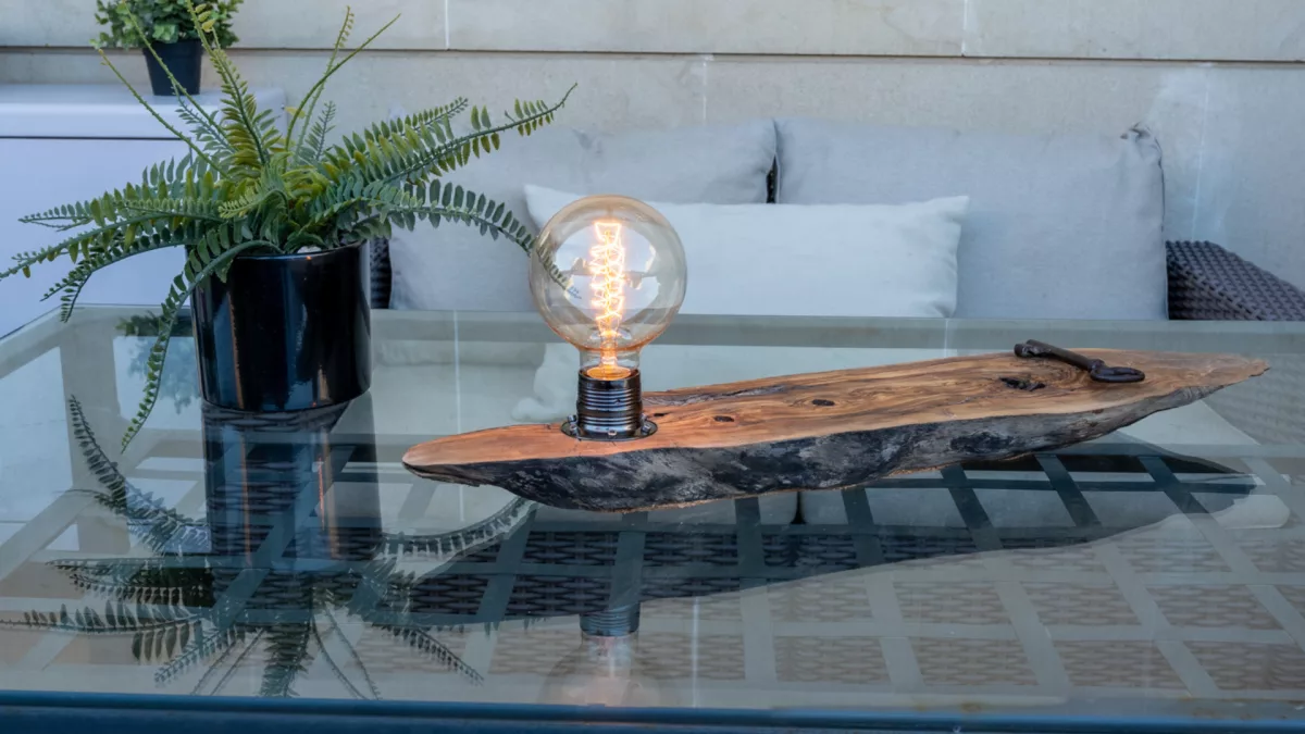 Lámpara decorativa con base de madera natural alargada y bombilla vintage encendida, sobre una mesa de cristal en un ambiente exterior.
